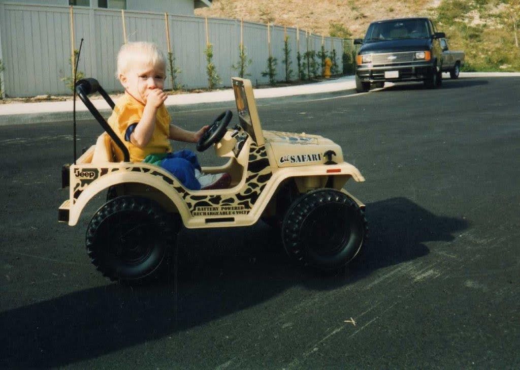 A toddler in a yellow toy Jeep on a driveway